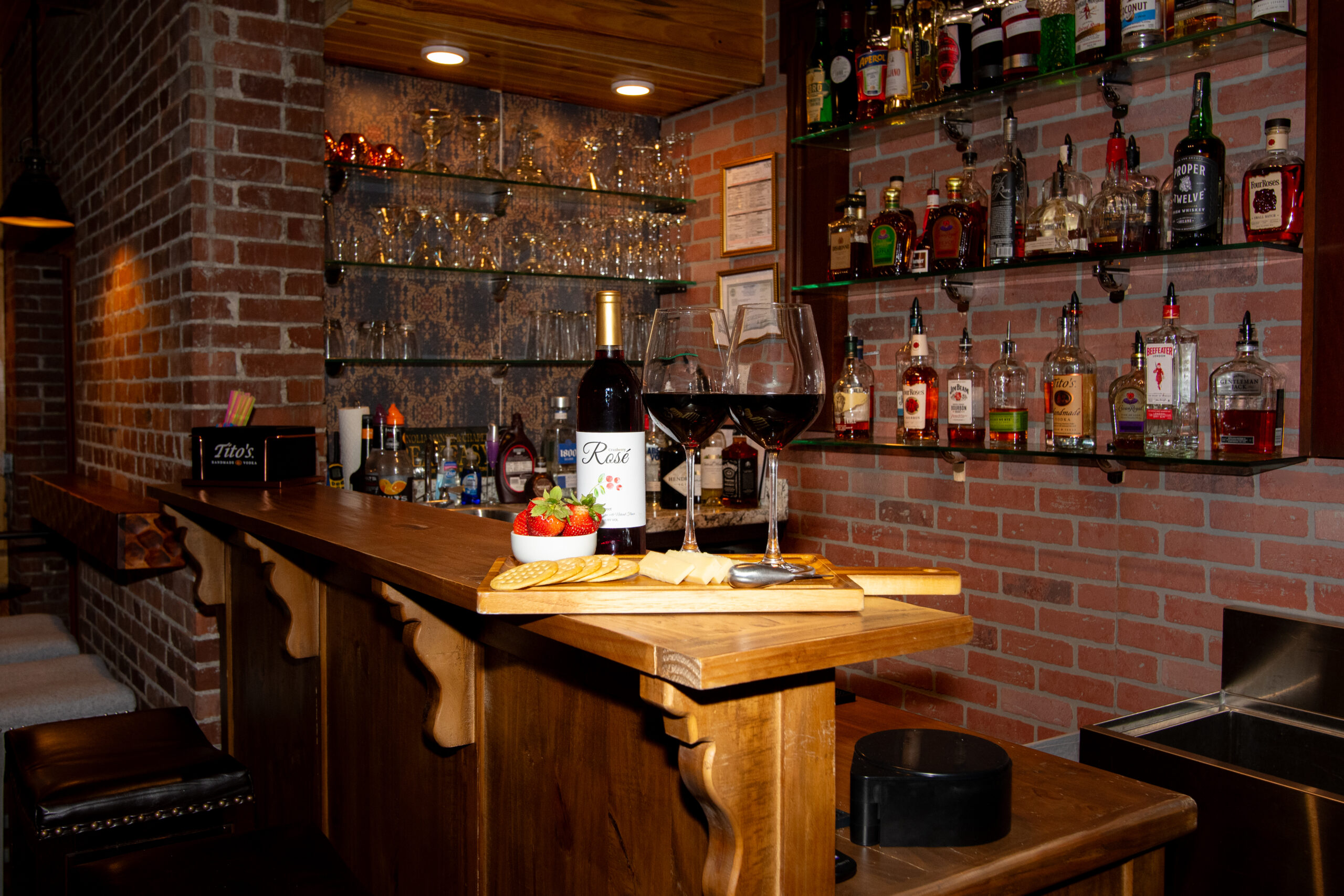 Two glasses of red wine on a wooden cheese board displayed with crackers, cheese, a white bowl of strawberries, and a bottle of sweet Cranberry Rosé wine
